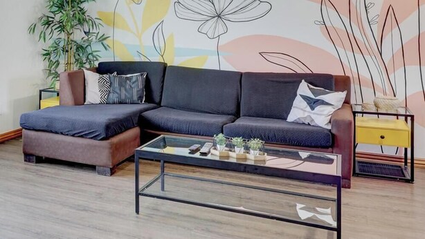 Una sala de estar moderna con un sofá modular, una mesa de centro de vidrio con plantas, una mesita auxiliar y una pared decorativa con diseños florales abstractos.