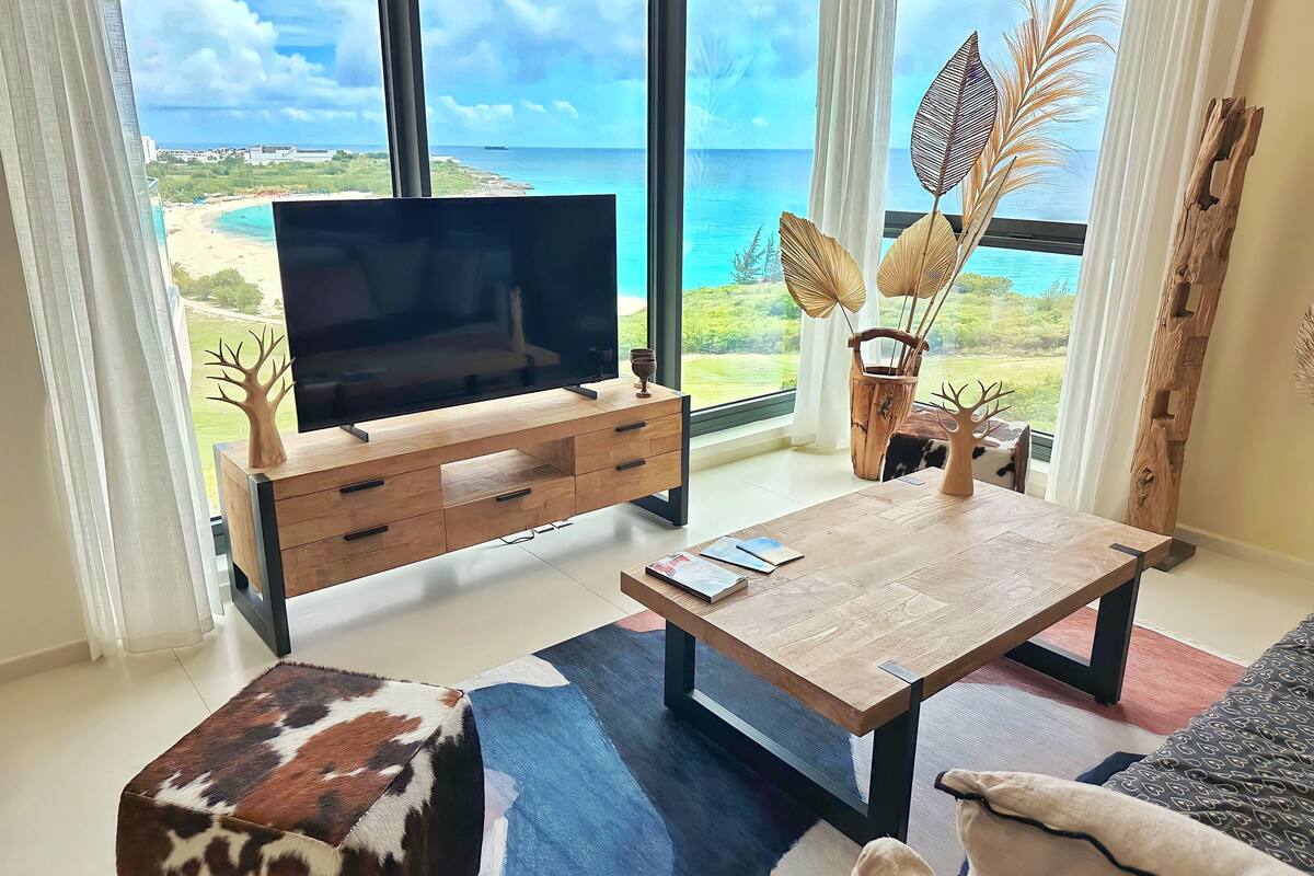 Un salón con televisor sobre un mueble de madera, una mesa de centro y vista al océano.