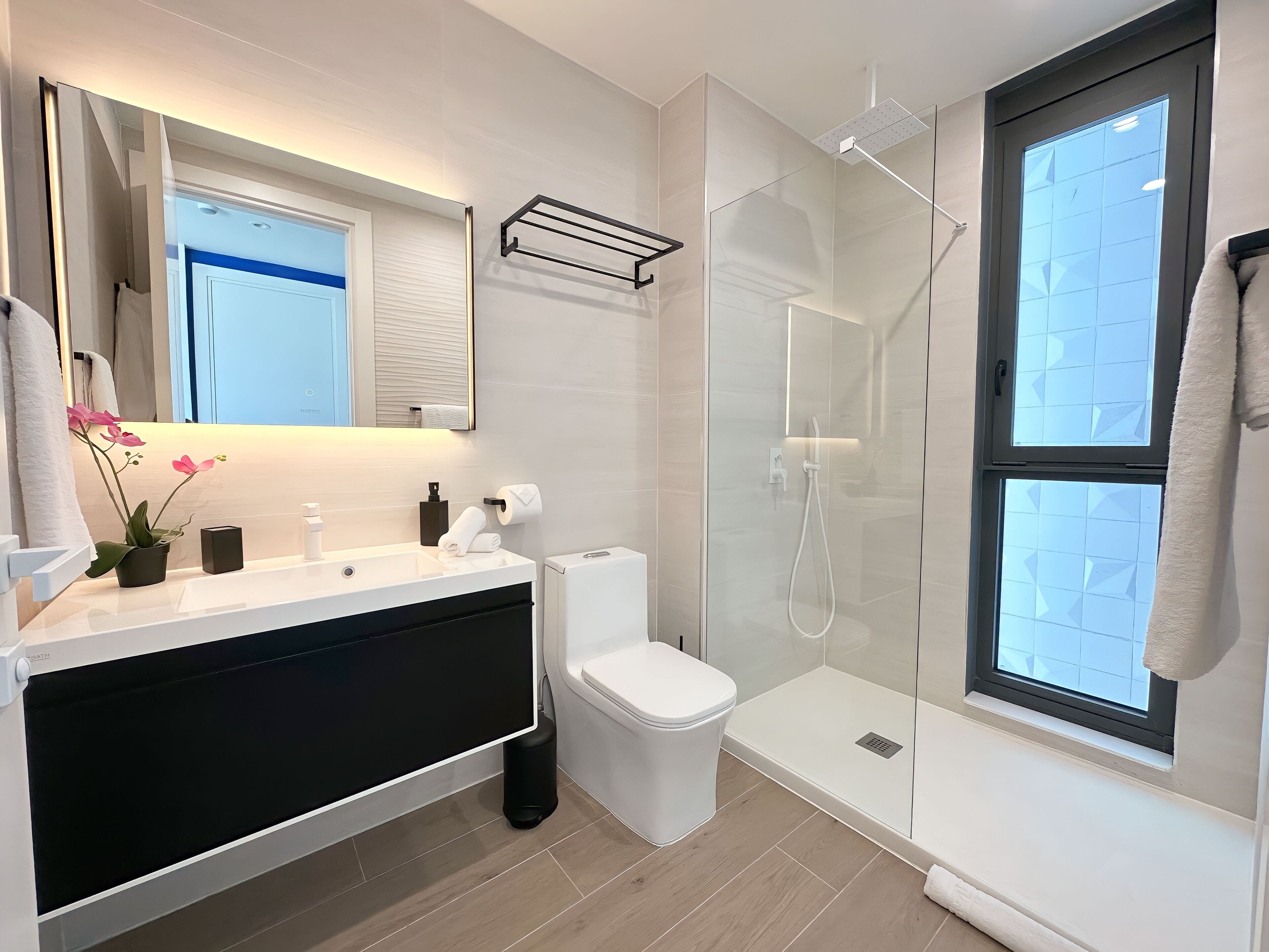 A modern bathroom with a glass shower enclosure, a white toilet, and a large window.