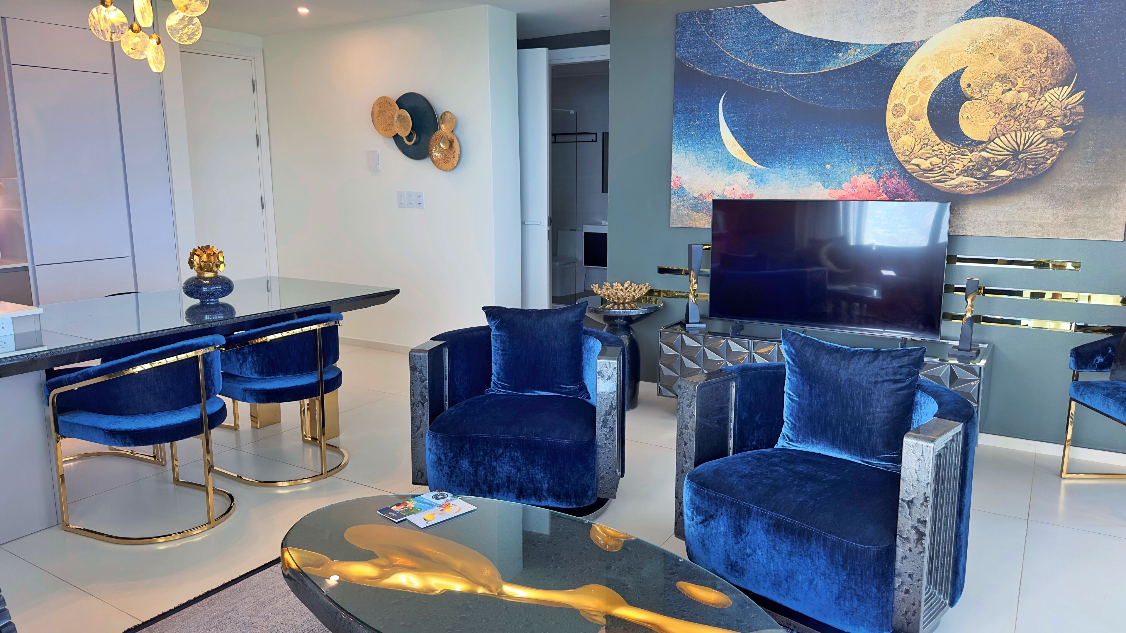 Una sala de estar moderna con televisor, sillones de terciopelo azul, una mesa de centro de vidrio y una obra de arte con luna y estrellas.