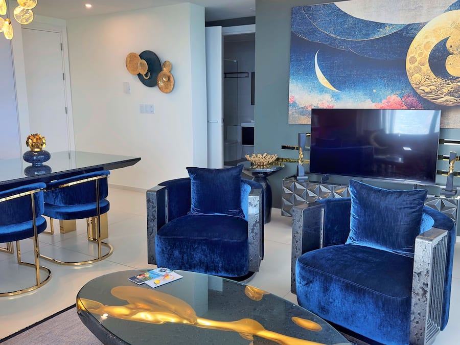 Una sala de estar moderna con televisor, sillones de terciopelo azul, una mesa de centro de vidrio y una obra de arte con luna y estrellas.