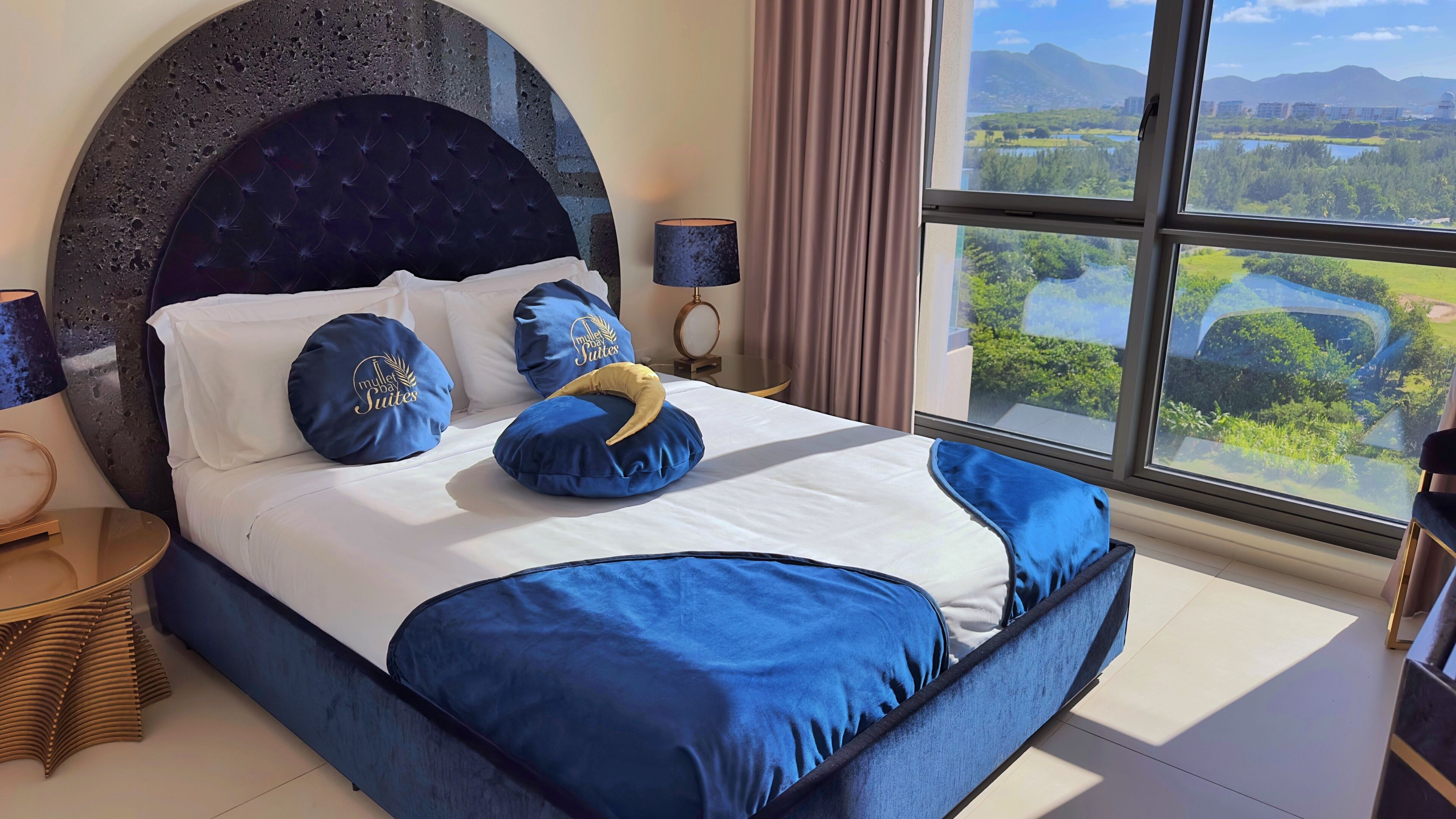 Una cama con cabecera oscura, ropa de cama blanca y almohadas azules con texto. Se ve una ventana con vistas a zonas verdes y edificios.