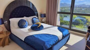 Una cama con cabecera oscura, ropa de cama blanca y almohadas azules con texto. Se ve una ventana con vistas a zonas verdes y edificios.
