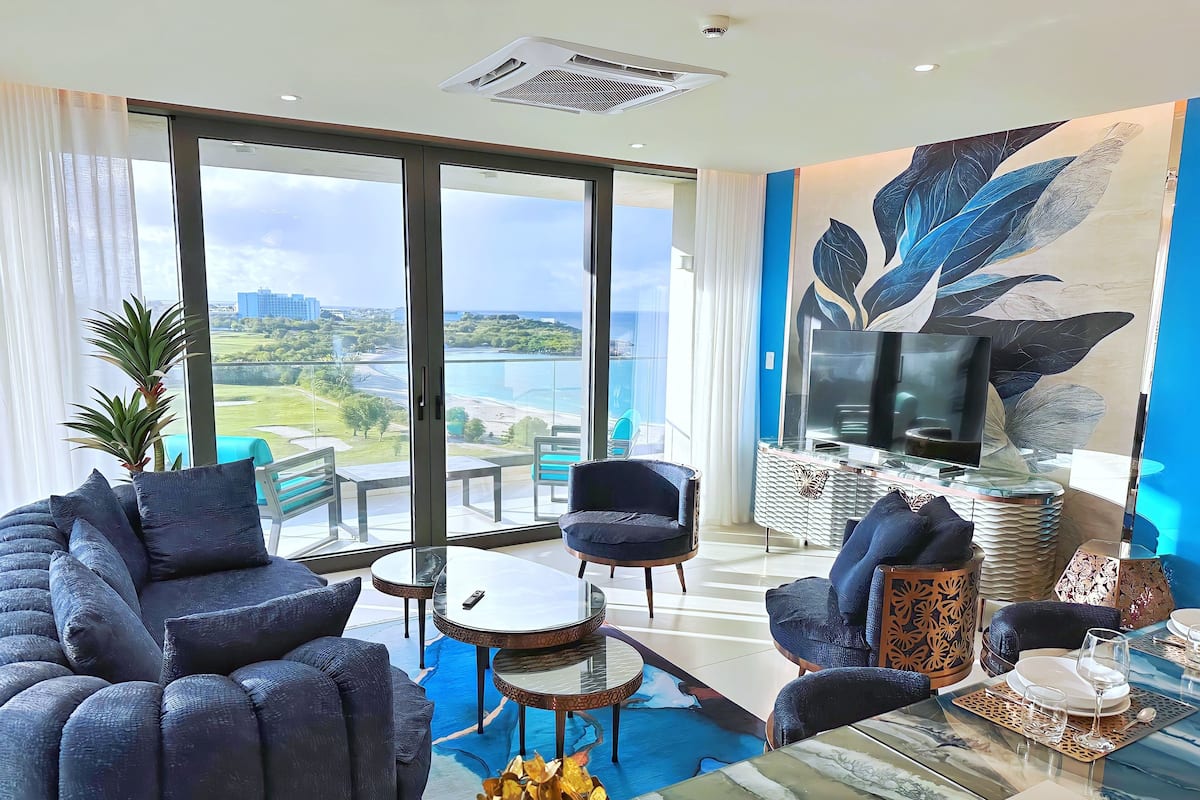 Una sala de estar moderna con un sofá grande, una mesa de centro y un televisor sobre un mueble bajo. La habitación tiene ventanas amplias con vistas a un paisaje urbano y zonas verdes.