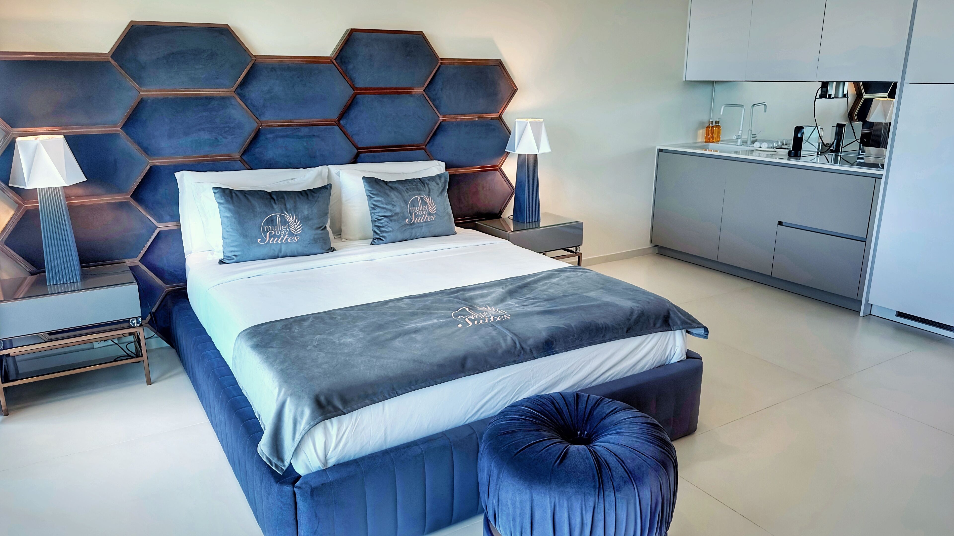 Un dormitorio moderno con un cabecero estampado con motivos geométricos, una cama grande con almohadas a juego, mesitas de noche con lámparas y una zona de kitchenette.