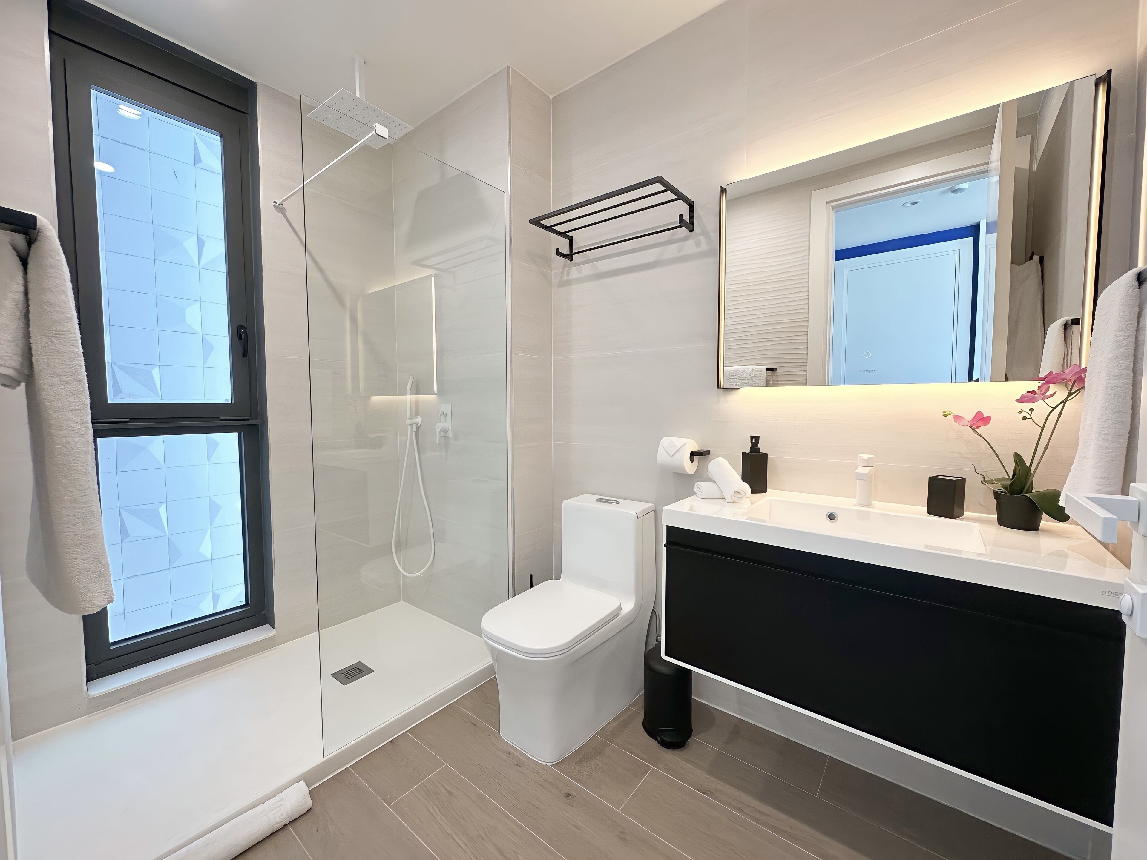 Un baño moderno con ducha envuelta en vidrio, inodoro, lavamanos con espejo y ventana.