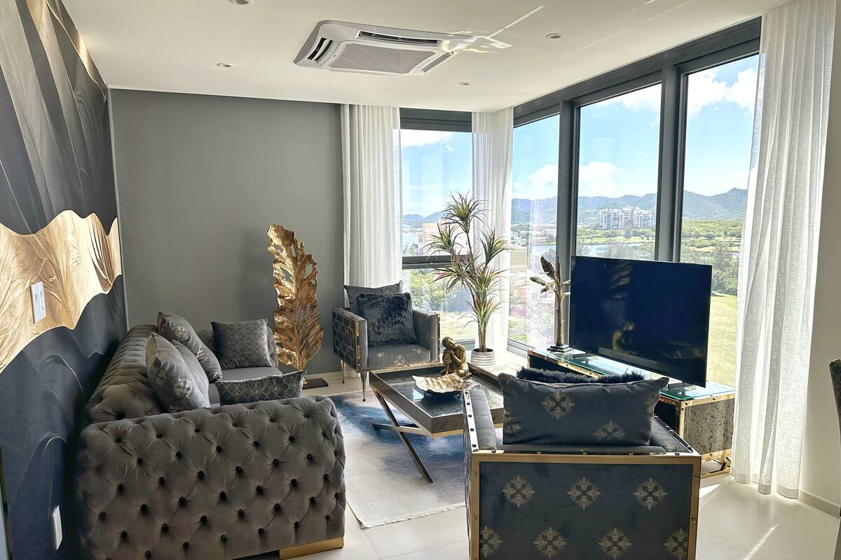 Una sala de estar moderna con un sofá gris, una mesa de centro y un televisor. Hay un ventanal con vistas a la ciudad y a las montañas.