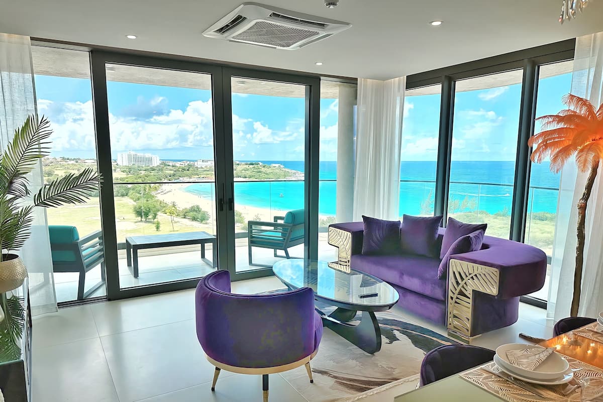 Una sala de estar moderna con ventanas amplias, un sofá púrpura y vista a la playa y al océano.