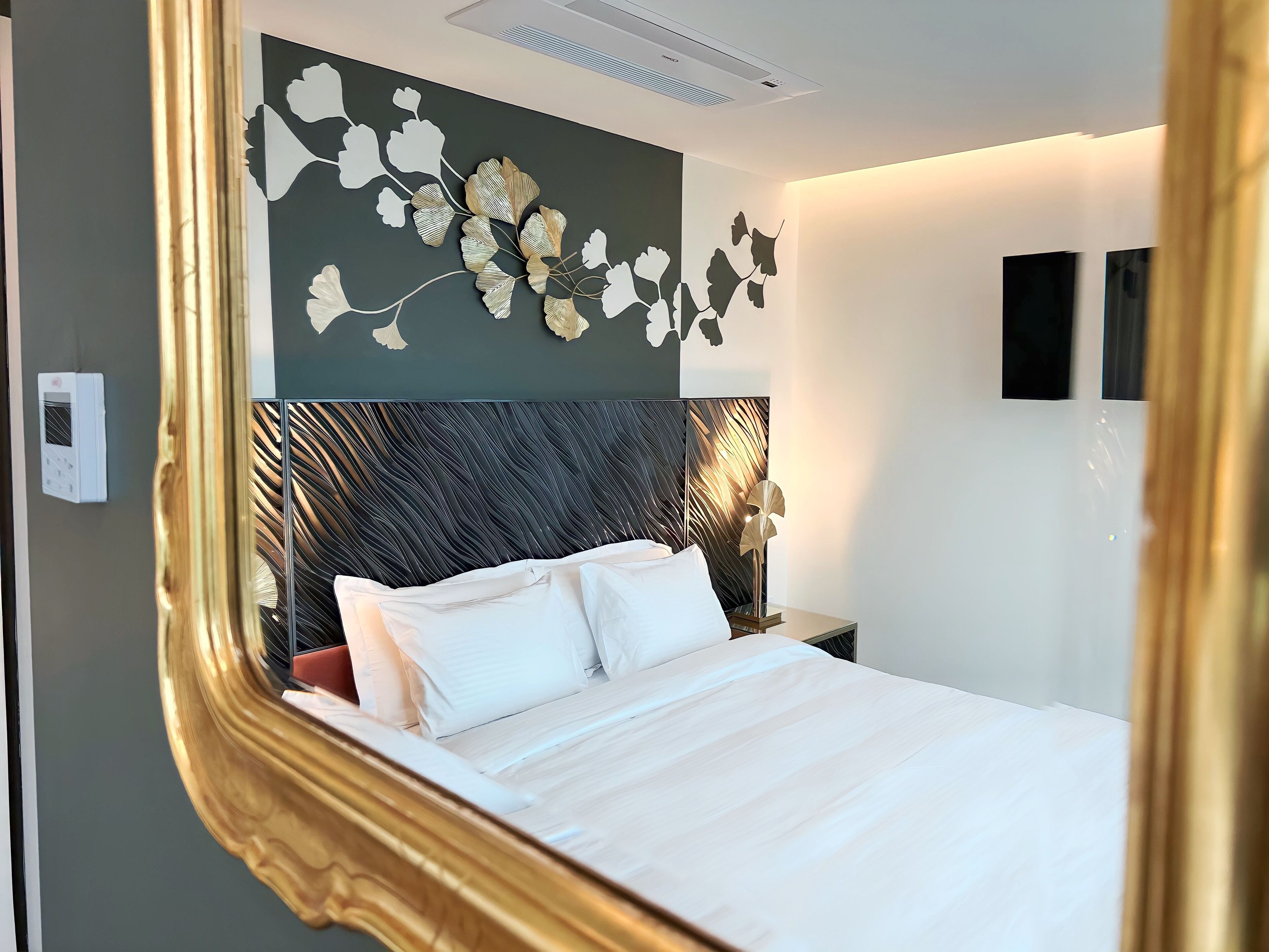 A modern hotel room with a large bed, a decorative wall panel, a wall-mounted air conditioner, and a bedside table with a lamp.
