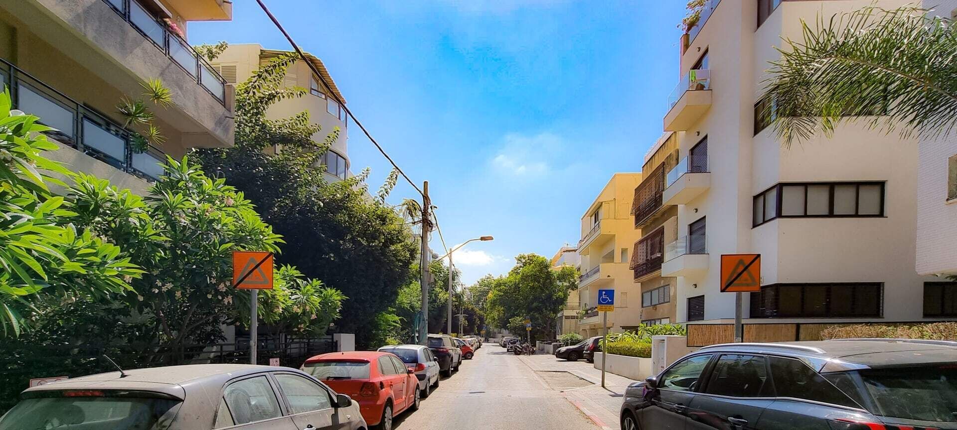 A narrow urban street lined with parked cars and multi-story residential buildings.