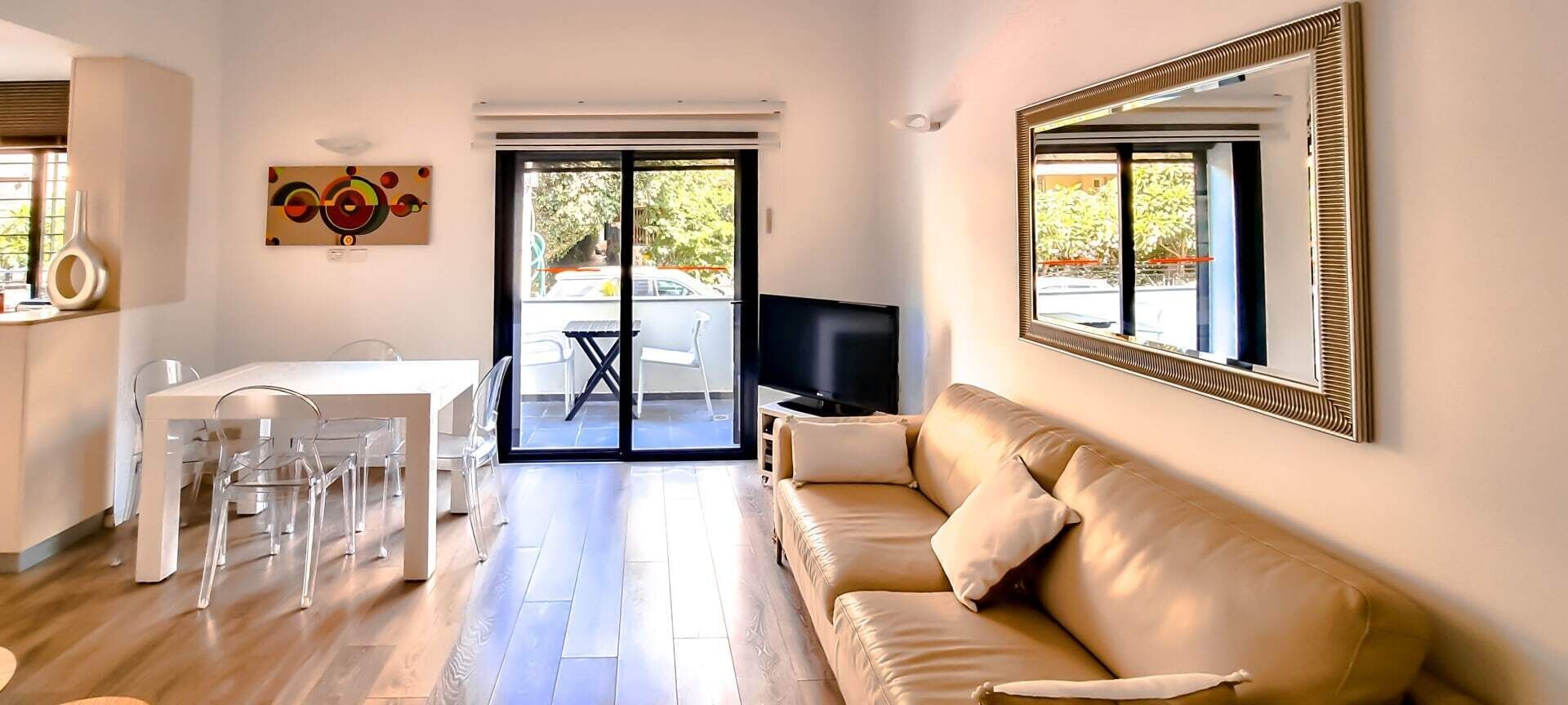 Una sala moderna con un sofá color beige, una mesa de comedor con sillas transparentes, un televisor de pantalla plana y un ventanal con vistas al exterior.
