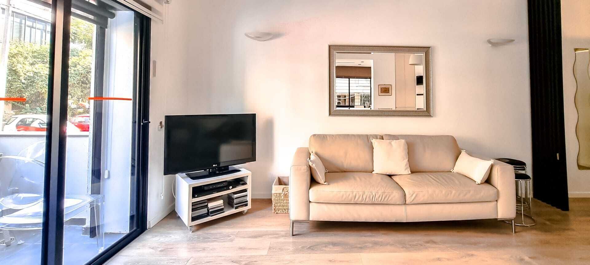 A modern living room with a flat-screen TV, a beige sofa, and a large mirror on the wall.