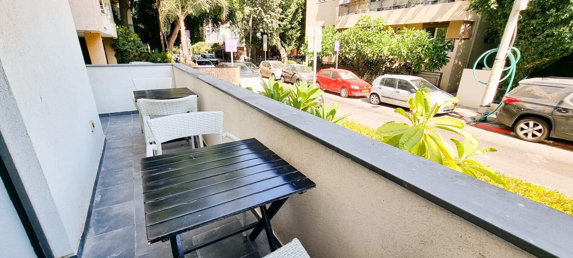 Un balcon avec une table noire et des chaises en osier blanc, donnant sur une rue bordée de voitures garées et d’espaces verts.