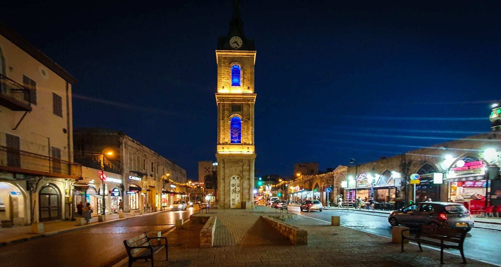 Ein historischer Glockenturm mit beleuchteten Fenstern, gelegen auf einem belebten Stadtplatz mit Geschäften und Cafés.