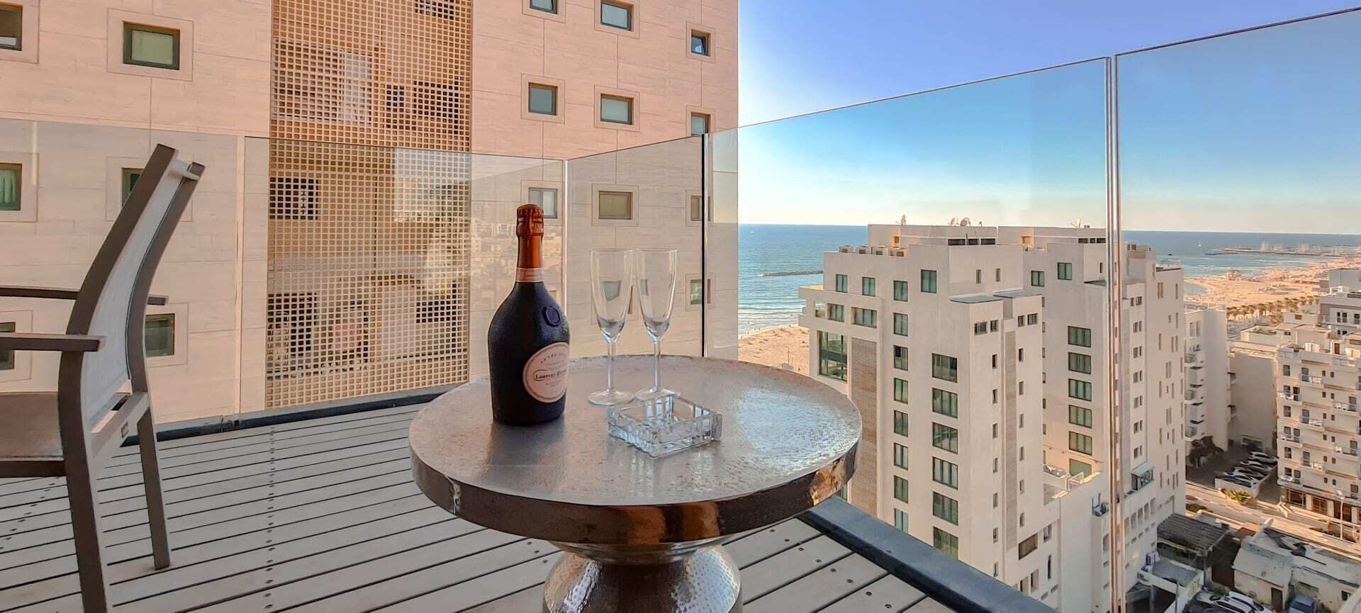 A balcony with a table, a bottle of wine, and a glass. The balcony overlooks a cityscape and the sea.