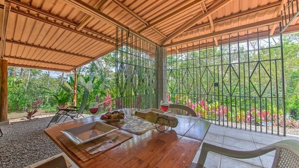 A covered outdoor dining area with a wooden table, chairs, and a view of greenery through metal bars.