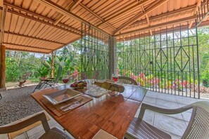 A covered outdoor dining area with a wooden table, chairs, and a view of greenery through metal bars.