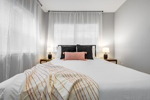A bedroom with a large bed, two bedside lamps, sheer curtains, and a window with blinds.