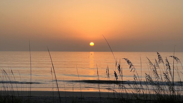 Eine Strandansicht mit Sonnenuntergang über dem Meer, ruhigem Wasser und hohem Gras im Vordergrund.