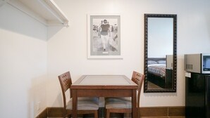 A small room with a dining table and chairs, a framed photograph on the wall, and a mirror reflecting a bed.