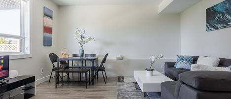 A modern living room with a grey sofa, a coffee table, a dining area with a table and chairs, and a TV on a stand.