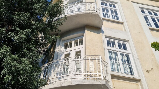 Una esquina de un edificio con balcón, ventanas y un árbol.