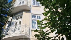 Un edificio de varios pisos con un balcón en el segundo nivel, ventanas amplias y un árbol en primer plano.