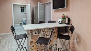 Una habitación con una mesa blanca, sillas negras, un televisor en la pared y una puerta que da a otra habitación.