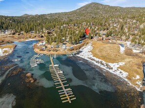 An aerial view of a riverside area with a wooden dock extending into the water, surrounded by residential buildings and a forested hillside. - North Shore Lakefront (Fawnskin)