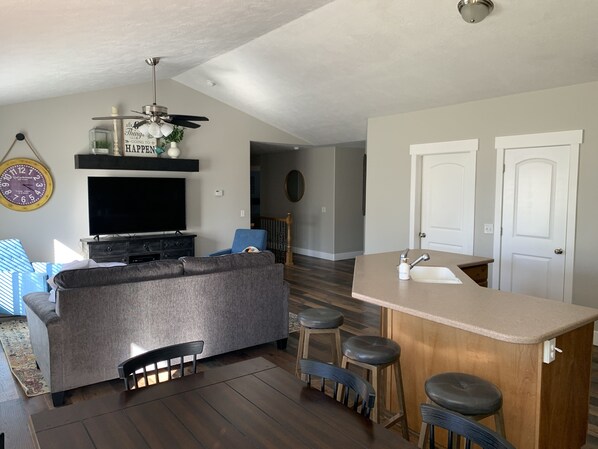 A modern living room with a gray sectional sofa, a flat-screen TV, a ceiling fan, and a wooden bar area with stools.