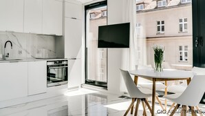 Una cocina moderna con armarios blancos, salpicadero de mármol y horno empotrado. Hay un ventanal con vistas a edificios y un televisor de pantalla plana montado en la pared.