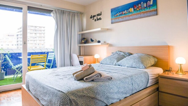 Ein Schlafzimmer mit Bett, Schreibtisch, Stuhl, Fenster mit Blick auf Gebäude und einem Bild an der Wand.