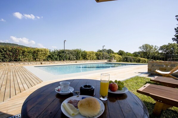 A wooden deck with a table set for breakfast, overlooking a swimming pool and lush greenery.