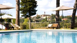 A swimming pool with lounge chairs and umbrellas, surrounded by trees and a view of buildings in the background.