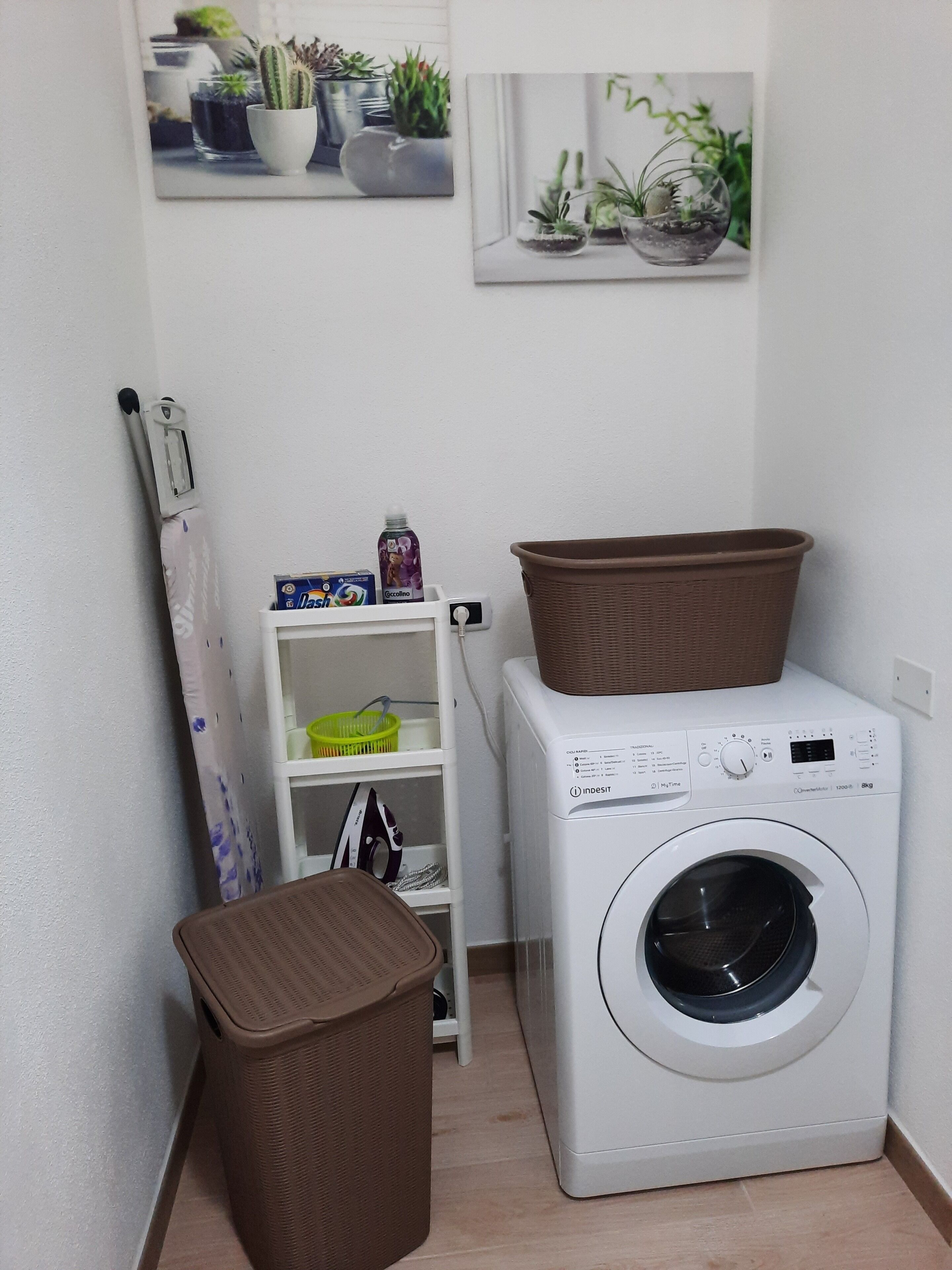 Een wasserette met een wasmachine, een mandje bovenop, een prullenbak en een plank met een mand en een fles.