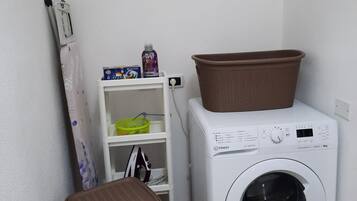 Een wasserette met een wasmachine, een mandje bovenop, een prullenbak en een plank met een mand en een fles.