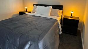 A neatly made bed with a dark gray comforter and white pillows, flanked by two bedside tables with lamps, in a room with a plain wall and carpeted floor. - Sydney Blue mountain Retreat (Kingswood)
