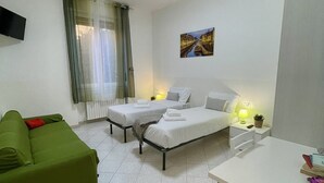 Una habitación pequeña y limpia con dos camas individuales, un sofá verde, una mesa blanca y un cuadro en la pared.