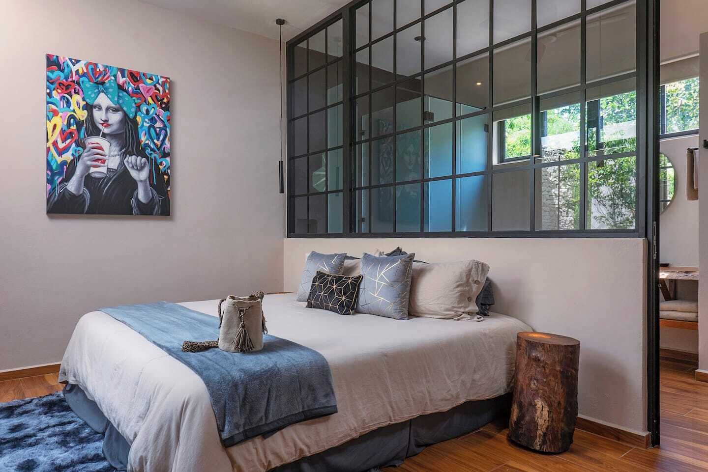 A modern bedroom with a large window, a bed with a blue blanket, and a painting on the wall.
