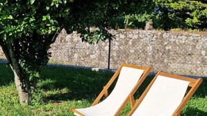 Deux chaises longues en bois, dotées d’assises et de dossiers en toile blanche, sont placées sur une pelouse, avec un mur de pierre et des arbres en arrière-plan.