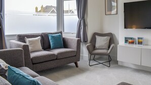 A modern living room with a grey sofa, a grey armchair, and a white cabinet. There is a large window with curtains, a flat-screen TV mounted on the wall, and a framed picture above the TV. - A beautifully presented, spacious holiday home. (Newquay)