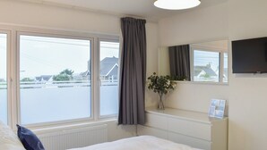 A hotel room with a large bed, a bedside table with a vase of flowers, a flat-screen TV mounted on the wall, and a window with curtains. - A beautifully presented, spacious holiday home. (Newquay)