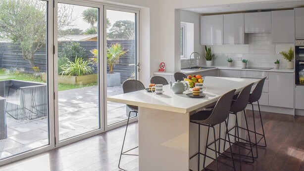 A modern kitchen with white cabinets, a central island, and a dining area with chairs. - A beautifully presented, spacious holiday home. (Newquay)