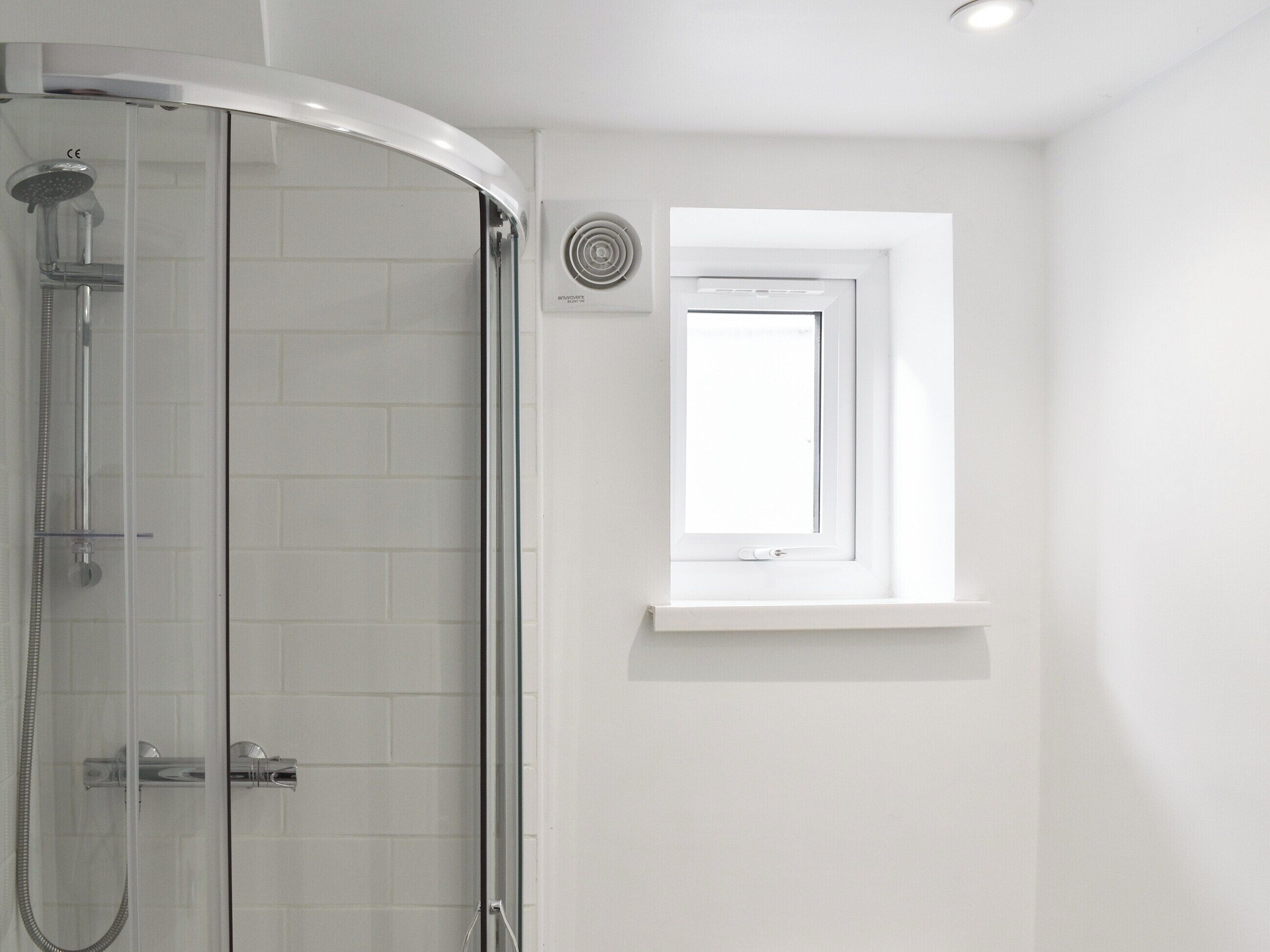 Eine Dusche mit Glastür, ein Fenster mit weißem Rahmen und eine Wand-Klimaanlage.