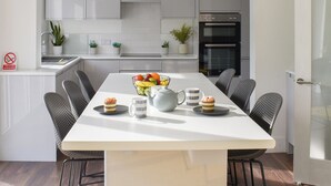 A modern kitchen with white cabinets, a built-in oven, and a dining area with a white table and chairs. - A beautifully presented, spacious holiday home. (Newquay)
