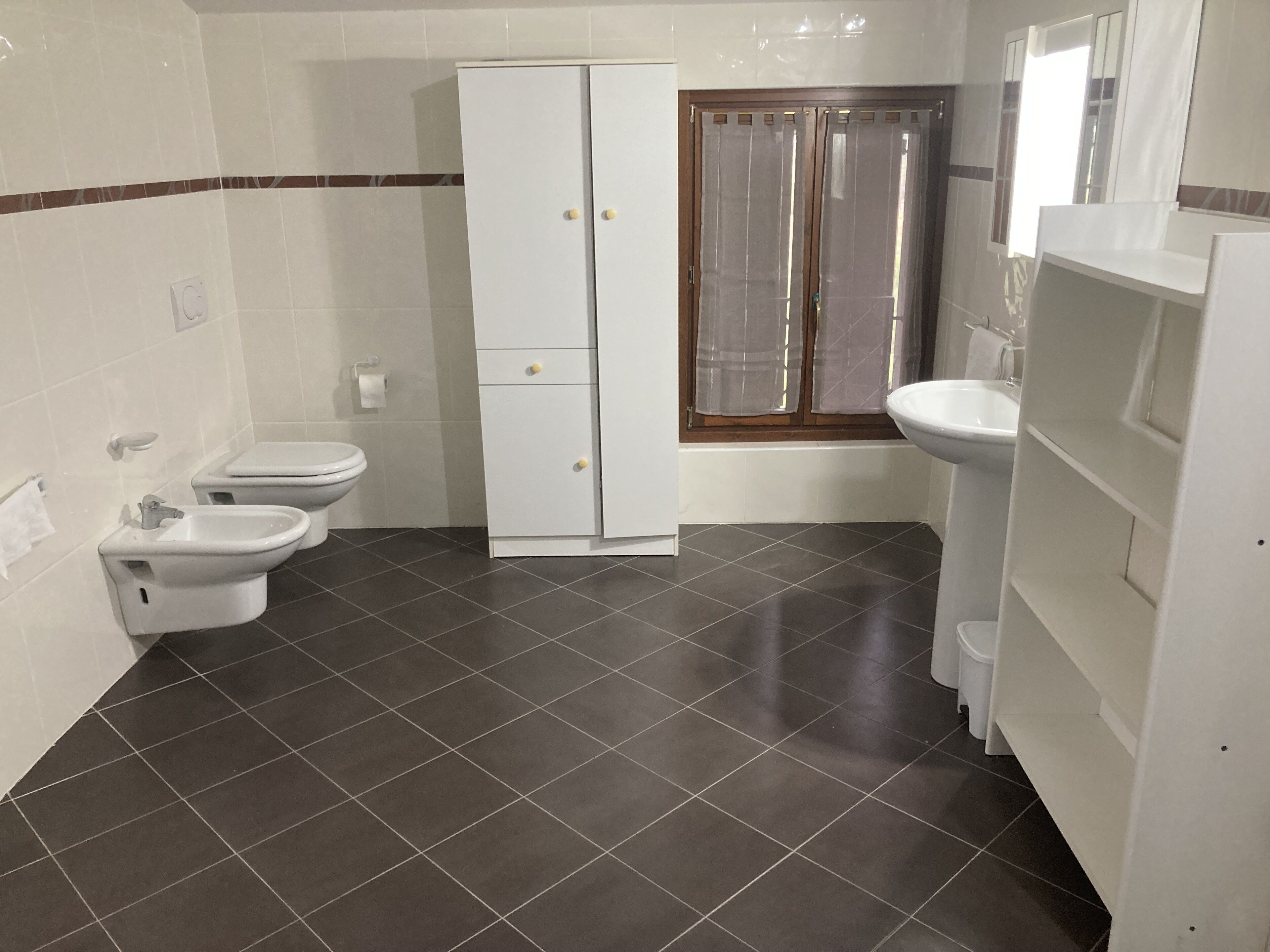 Une salle de bain avec deux toilettes, un bidet, un meuble, un lavabo et une fenêtre avec des rideaux.