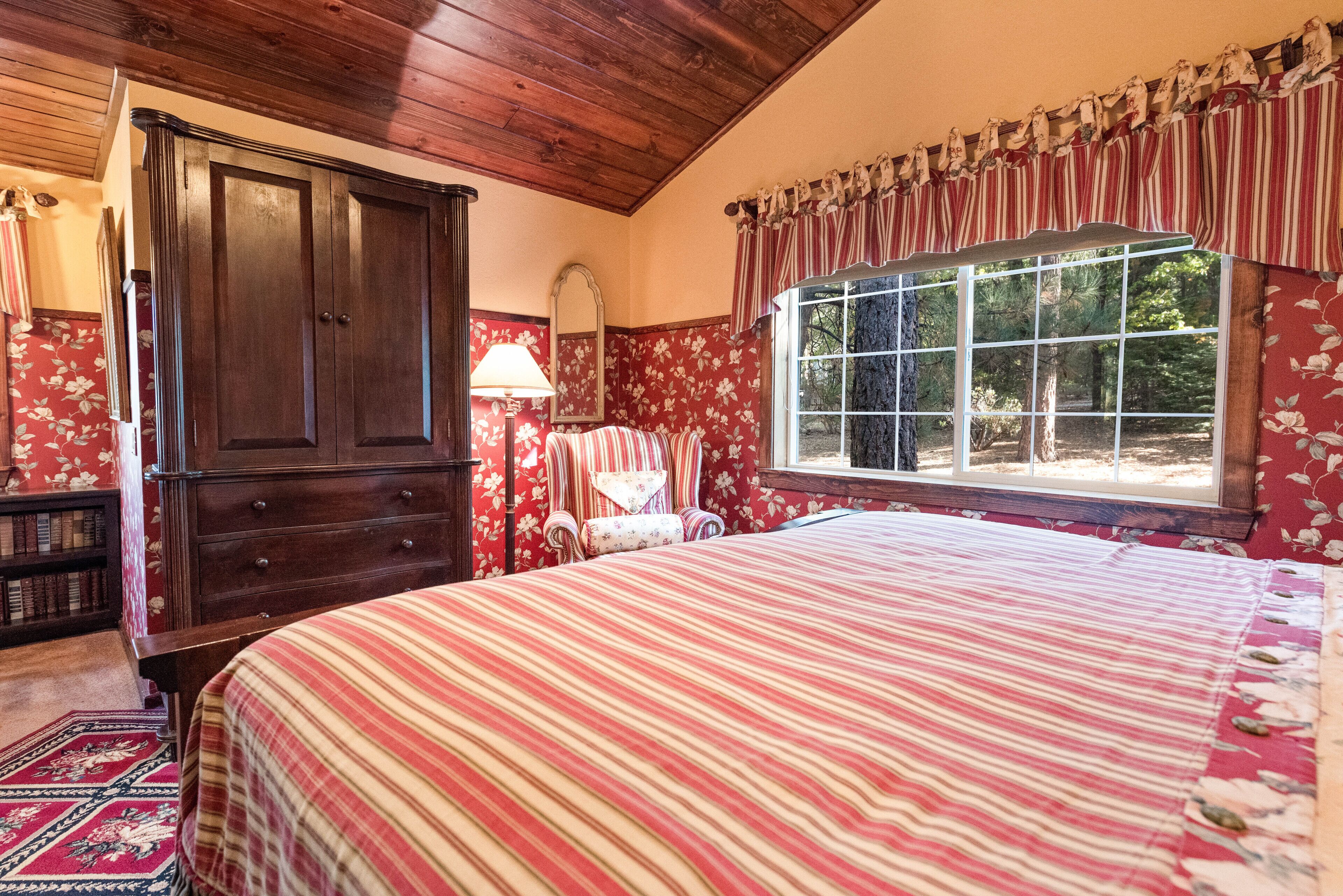 A bedroom with a large bed, wooden wardrobe, floral wallpaper, and a window with curtains.