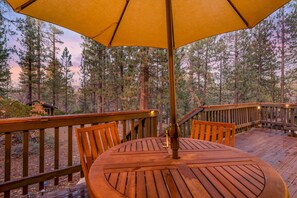 Eine Holzterrasse mit rundem Tisch und Stühlen, umgeben von einem Geländer, mit Blick auf ein bewaldetes Gebiet.