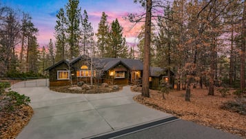 A log cabin with a concrete driveway, surrounded by tall pine trees and a sunset sky.