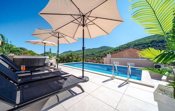 Área da piscina com várias espreguiçadeiras e guarda-sóis, uma banheira de hidromassagem e vista para uma paisagem montanhosa.